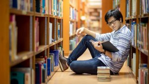 Asian man sitting on the floor between book stacks in a library reading a book