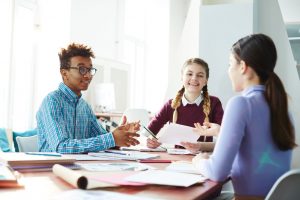 A group of three students sitting around a table with lots of papers and talking wtih each other